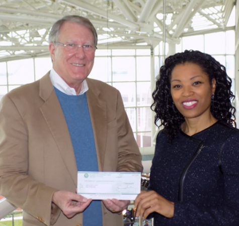 A woman with dark curly hair wearing a dark shirt hands a check to a man wearing a blue shirt and tan jacket.