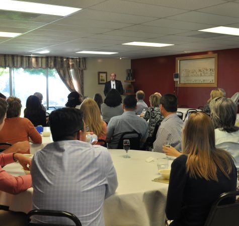 A group of people sitting around tables listen to a speaker at the front of the room.