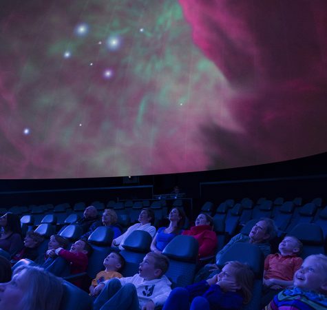 An audience sits inside a planetarium auditorium while looking up at a projection of the galaxies.