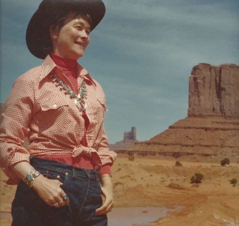 A woman wearing a red button-down shirt, jeans, and a cowboy hat standing outside smiles at something off camera.